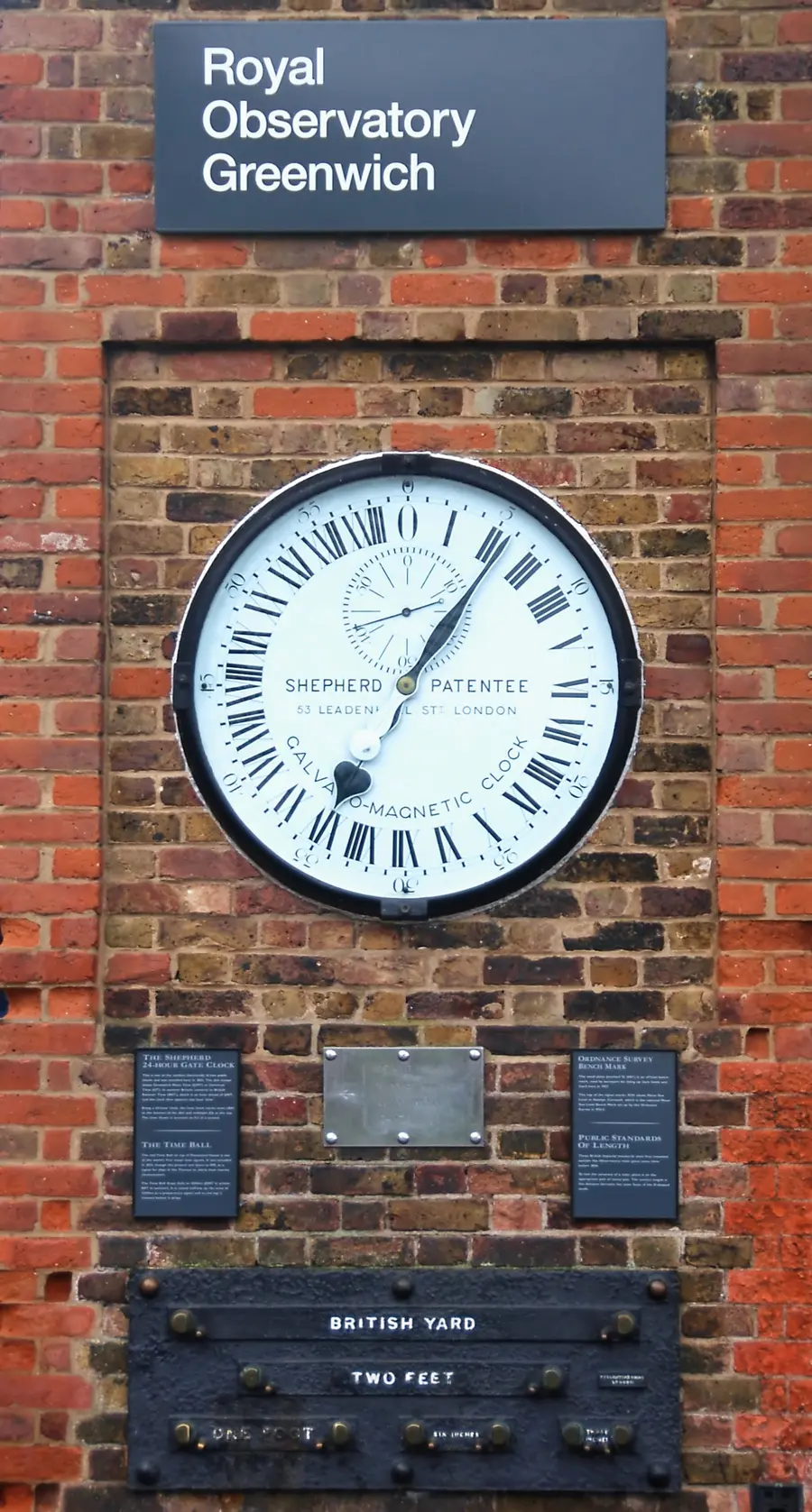 A large 24-hour clock face set into a red brick wall beneath a sign reading Royal Observatory Greenwich